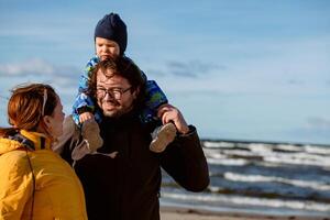 A happy family strolls along a chilly autumn beach, with their young son joyfully running ahead, creating lasting memories as the crisp wind and soothing waves embrace them in a moment of pure photo