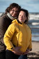 A couple stands embraced on a sandy beach on a chilly autumn day, the serene ocean stretching behind them as they share a quiet moment of love and warmth amidst the cool breeze and peaceful photo