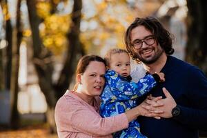 On a sunny autumn day in the park, a mother and father lovingly embrace their young son, sharing moments of laughter and joy as they play together photo