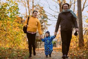 A loving couple strolls through a sunlit park with their young son, surrounded by the vibrant colors of autumn, enjoying a joyful and peaceful family moment together. photo