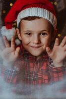 the joyful face of a boy in a Santa hat at a snow-covered window. Festive mood. photo