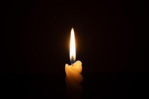 A single burning candle flame or light glowing on a spiral white candle on black or dark background on table in church for Christmas, funeral or memorial service with copy space photo