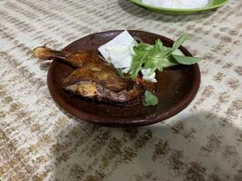 Ayam bakar Penyet is a fried chicken served with hot chili in a plate with fresh vegetables, slices of cucumber and basil leaves photo