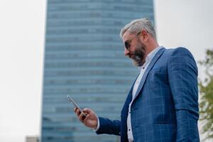 A handsome mature business man using mobile phone app texting outside of office in city with urban buildings in the background. photo