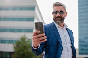A happy mature business man using a cell phone app texting outside of the office in the city with urban buildings in the background. photo