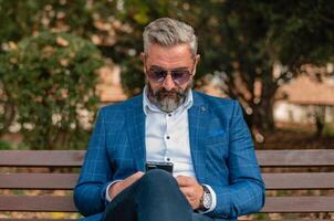 A hipster mature business man using mobile phone app texting while sitting on the park bench. photo