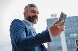 A happy mature business man using mobile phone app texting outside of office in city with urban buildings in the background. photo