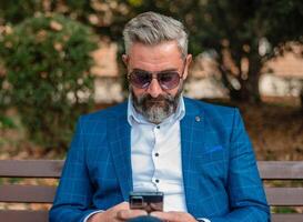 A hipster mature business man using mobile phone app texting while sitting on the park bench. photo