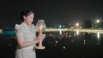 Bathed in the soft glow of lantern light, a woman stands in prayer beside a serene pond, her offering symbolizing gratitude and hope during the enchanting Loy Krathong festival. video