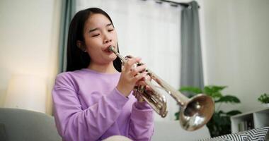Handheld low angle view shot, Relaxed Asian woman sitting on a sofa enjoying playing the trumpet in a cozy room at home. Practice and dedication to favorite musical instrument. video