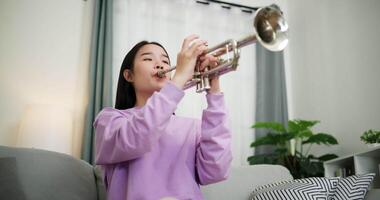 Handheld low angle view shot, Relaxed Asian woman sitting on a sofa enjoying playing the trumpet in a cozy room at home. Practice and dedication to favorite musical instrument. video