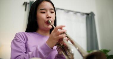 Handheld low angle view shot, Relaxed Asian woman sitting on a sofa enjoying playing the trumpet in a cozy room at home. Practice and dedication to favorite musical instrument. video