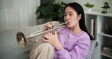 Handheld shot, Relaxed Asian woman sitting on a sofa enjoying playing the trumpet in a cozy room at home. Practice and dedication to favorite musical instrument. video