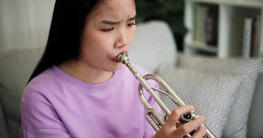 Handheld high angle view shot, Relaxed Asian woman sitting on a sofa enjoying playing the trumpet in a cozy room at home. Practice and dedication to favorite musical instrument. video