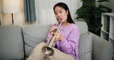 Handheld shot, Beautiful young woman in a light purple sweater sits on a sofa and is practicing the trumpet with determination in a cozy room at home. Weekend leisure activity. video