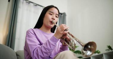 Handheld low angle view shot, Relaxed Asian woman sitting on a sofa enjoying playing the trumpet in a cozy room at home. Practice and dedication to favorite musical instrument. video