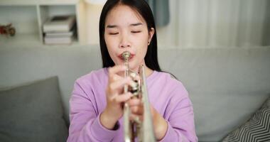 Handheld high angle view shot, Relaxed Asian woman sitting on a sofa enjoying playing the trumpet in a cozy room at home. Practice and dedication to favorite musical instrument. video