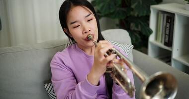 Handheld high angle view shot, Beautiful young woman in a light purple sweater sits on a sofa and is practicing the trumpet with determination in a cozy room at home. Weekend leisure activity. video