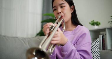 Handheld shot, Relaxed Asian woman sitting on a sofa enjoying playing the trumpet in a cozy room at home. Practice and dedication to favorite musical instrument. video