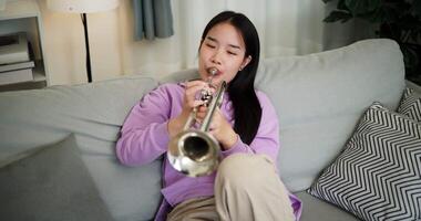 Handheld high angle view shot, Relaxed Asian woman sitting on a sofa enjoying playing the trumpet in a cozy room at home. Practice and dedication to favorite musical instrument. video