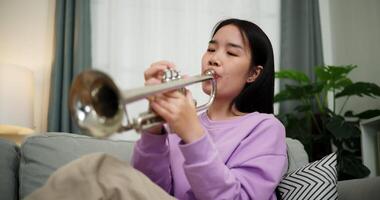 Handheld low angle view shot, Beautiful young woman in a light purple sweater sits on a sofa and is practicing the trumpet with determination in a cozy room at home. Weekend leisure activity. video