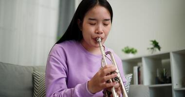 Handheld low angle view shot, Beautiful young woman in a light purple sweater sits on a sofa and is practicing the trumpet with determination in a cozy room at home. Weekend leisure activity. video
