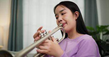 Handheld low angle view shot, Relaxed Asian woman sitting on a sofa enjoying playing the trumpet in a cozy room at home. Practice and dedication to favorite musical instrument. video