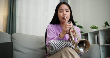 Handheld low angle view shot, Relaxed Asian woman sitting on a sofa enjoying playing the trumpet in a cozy room at home. Practice and dedication to favorite musical instrument. video