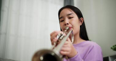Handheld low angle view shot, Relaxed Asian woman sitting on a sofa enjoying playing the trumpet in a cozy room at home. Practice and dedication to favorite musical instrument. video