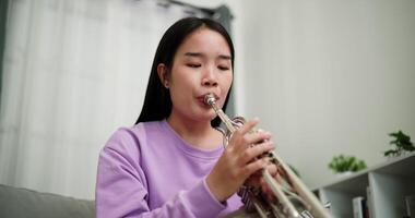 Handheld low angle view shot, Beautiful young woman in a light purple sweater sits on a sofa and is practicing the trumpet with determination in a cozy room at home. Weekend leisure activity. video