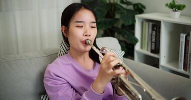 Handheld high angle view shot, Beautiful young woman in a light purple sweater sits on a sofa and is practicing the trumpet with determination in a cozy room at home. Weekend leisure activity. video