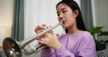 Handheld low angle view shot, Relaxed Asian woman sitting on a sofa enjoying playing the trumpet in a cozy room at home. Practice and dedication to favorite musical instrument. video