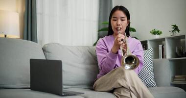 Relaxed young woman sitting on sofa and enjoying playing trumpet while looking at music notes on laptop in living room at home. Practice and dedication to favorite musical instrument. video