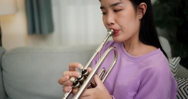 Handheld high angle view shot, Relaxed Asian woman sitting on a sofa enjoying playing the trumpet in a cozy room at home. Practice and dedication to favorite musical instrument. video