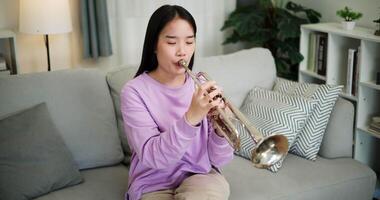 Handheld high angle view shot, Relaxed Asian woman sitting on a sofa enjoying playing the trumpet in a cozy room at home. Practice and dedication to favorite musical instrument. video