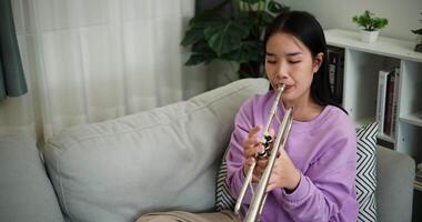 Handheld high angle view shot, Relaxed Asian woman sitting on a sofa enjoying playing the trumpet in a cozy room at home. Practice and dedication to favorite musical instrument. video