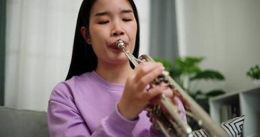 Handheld low angle view shot, Beautiful young woman in a light purple sweater sits on a sofa and is practicing the trumpet with determination in a cozy room at home. Weekend leisure activity. video