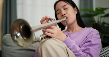 Handheld selective focus shot, Beautiful young woman in a light purple sweater sits on a sofa and is practicing the trumpet with determination in a cozy room at home. Weekend leisure activity. video
