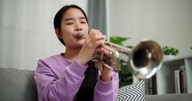 Handheld low angle view shot, Beautiful young woman in a light purple sweater sits on a sofa and is practicing the trumpet with determination in a cozy room at home. Weekend leisure activity. video
