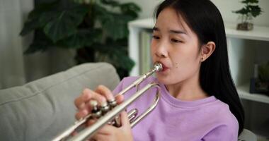 Handheld high angle view shot, Relaxed Asian woman sitting on a sofa enjoying playing the trumpet in a cozy room at home. Practice and dedication to favorite musical instrument. video