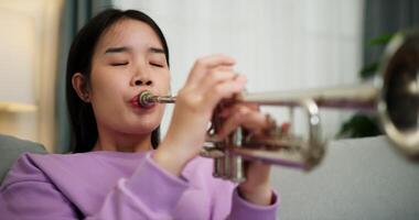 Handheld low angle view shot, Relaxed Asian woman sitting on a sofa enjoying playing the trumpet in a cozy room at home. Practice and dedication to favorite musical instrument. video