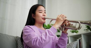 Handheld low angle view shot, Beautiful young woman in a light purple sweater sits on a sofa and is practicing the trumpet with determination in a cozy room at home. Weekend leisure activity. video