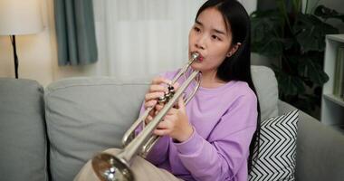 Handheld high angle view shot, Beautiful young woman in a light purple sweater sits on a sofa and is practicing the trumpet with determination in a cozy room at home. Weekend leisure activity. video