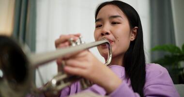Handheld low angle view shot, Relaxed Asian woman sitting on a sofa enjoying playing the trumpet in a cozy room at home. Practice and dedication to favorite musical instrument. video