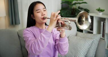 Handheld high angle view shot, Relaxed Asian woman sitting on a sofa enjoying playing the trumpet in a cozy room at home. Practice and dedication to favorite musical instrument. video