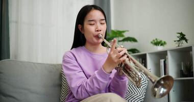 Handheld low angle view shot, Beautiful young woman in a light purple sweater sits on a sofa and is practicing the trumpet with determination in a cozy room at home. Weekend leisure activity. video