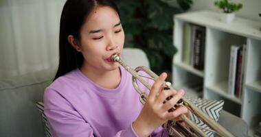 Handheld high angle view shot, Relaxed Asian woman sitting on a sofa enjoying playing the trumpet in a cozy room at home. Practice and dedication to favorite musical instrument. video