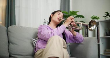 Handheld low angle view shot, Beautiful young woman in a light purple sweater sits on a sofa and is practicing the trumpet with determination in a cozy room at home. Weekend leisure activity. video