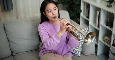 Handheld high angle view shot, Beautiful young woman in a light purple sweater sits on a sofa and is practicing the trumpet with determination in a cozy room at home. Weekend leisure activity. video
