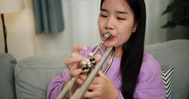 Handheld high angle view shot, Beautiful young woman in a light purple sweater sits on a sofa and is practicing the trumpet with determination in a cozy room at home. Weekend leisure activity. video